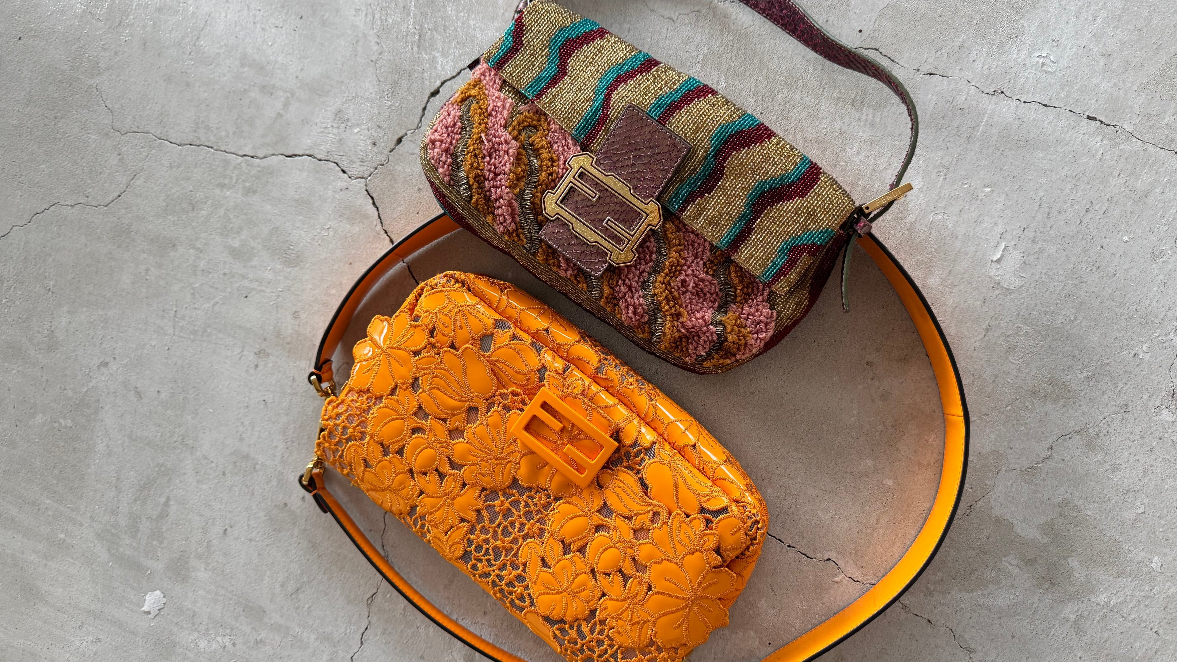 Two vintage Baguette shoulder bags from Fendi displayed on a gray concrete floor. The left bag is made of vibrant orange embossed leather with a matching orange strap and a small orange clasp. The right one is fully beaded and embroidered in multicolour design.